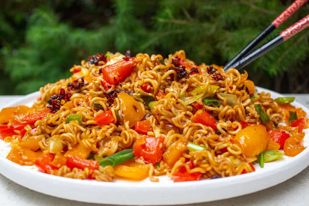 A plate of spicy stir-fried noodles with colorful bell peppers, chopped vegetables, and a dark chili sauce on top. Chopsticks are poised above the dish, and a backdrop of greenery is visible in the background.