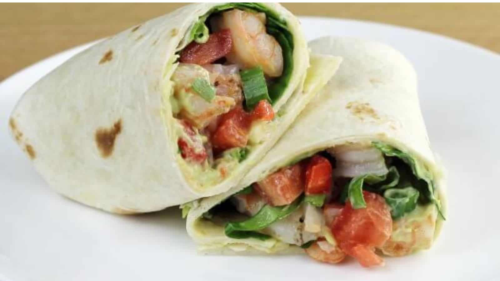 A close-up of two halves of a wrap on a white plate. The wrap is filled with shrimp, lettuce, red peppers, tomatoes, and green onions, all inside a soft tortilla. The ingredients are vibrant and freshly prepared.