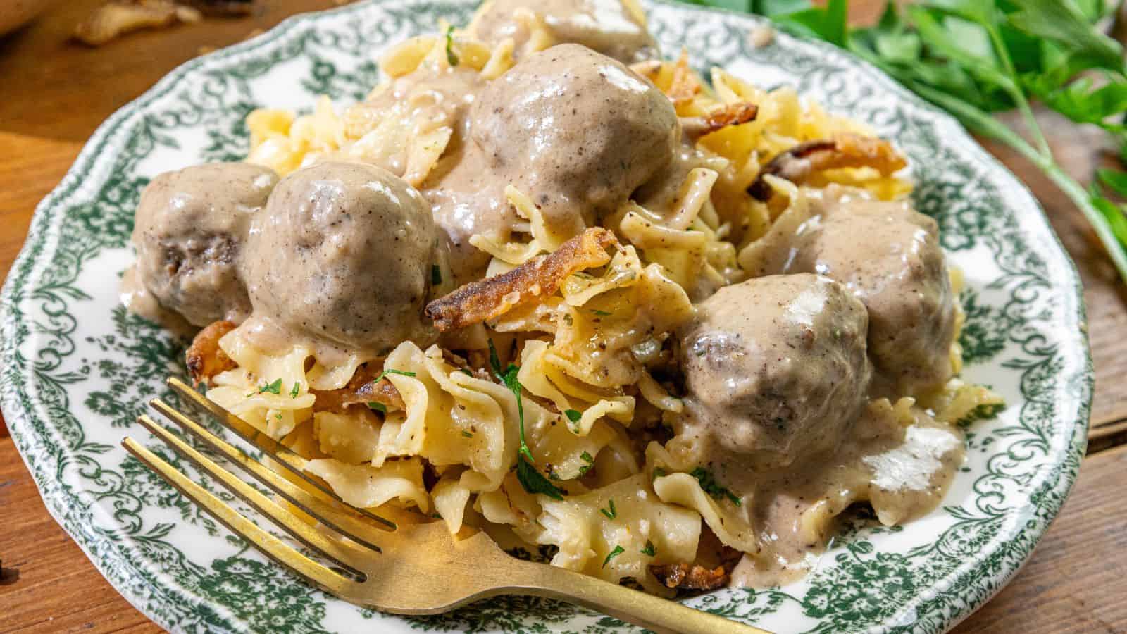 A plate of egg noodles with meatballs, creamy gravy, and caramelized onions, served with a gold fork on the side.