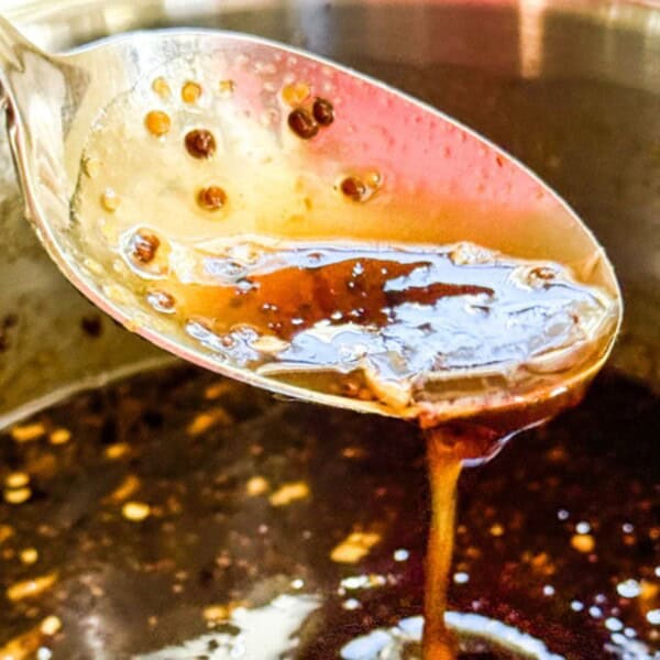 A spoon drizzles a thick, brown sauce with visible spices back into a metal pot.