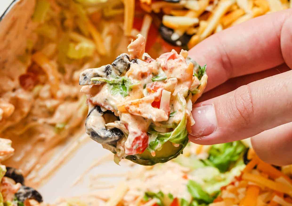A close-up of a hand holding a chip topped with a creamy dip made of sliced olives, shredded cheese, diced tomatoes, and lettuce. The dip is in a dish in the background, with more chips nearby.