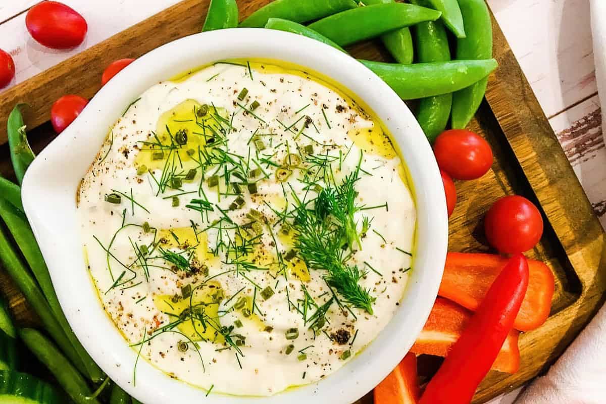 A white bowl filled with creamy dip, drizzled with olive oil and garnished with fresh herbs. It's surrounded by cherry tomatoes, snap peas, and sliced red bell peppers on a wooden serving board.