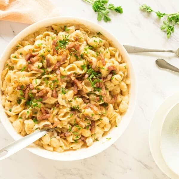 Creamy pasta with chopped bacon and herbs in a bowl, on marble with two forks, beige napkin, and an empty bowl nearby.