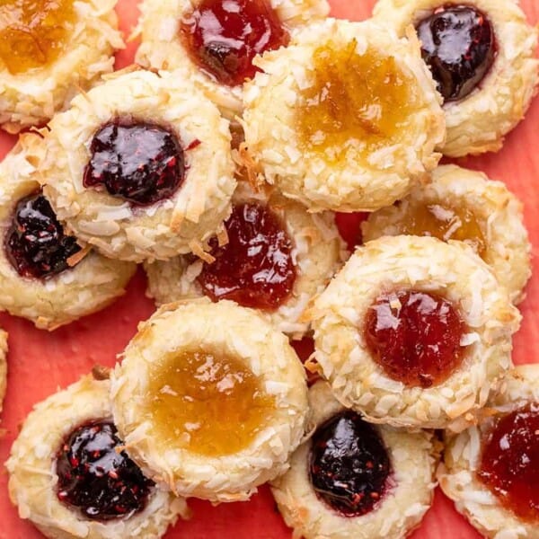 Coconut flake thumbprint cookies with different fruit jams are arranged among other Christmas cookies on a pink background.