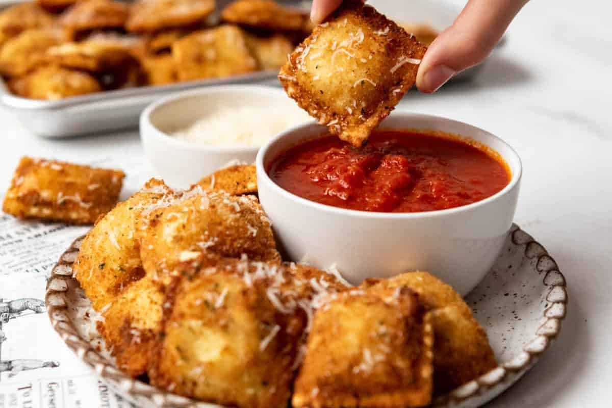 A plate of crispy fried ravioli, sprinkled with grated cheese, is served alongside a bowl of marinara sauce. A hand is seen dipping a ravioli into the sauce, with more ravioli on a tray in the background.