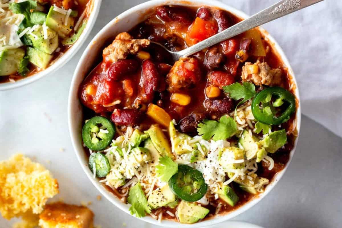 A bowl of chili topped with sliced jalapeños, diced avocado, shredded cheese, and cilantro. A spoon rests in the bowl, and cornbread is visible on the side. The dish contains beans, tomatoes, and chunks of meat.