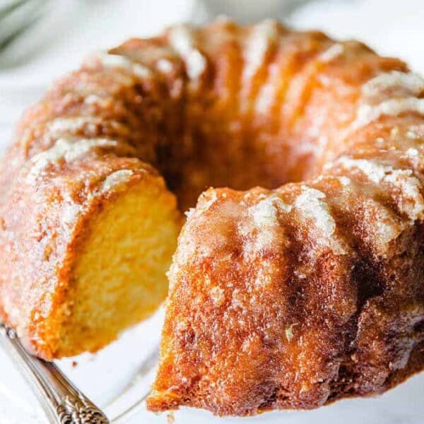 A golden bundt cake with a slice missing sits on a clear glass cake stand next to a metal cake server.