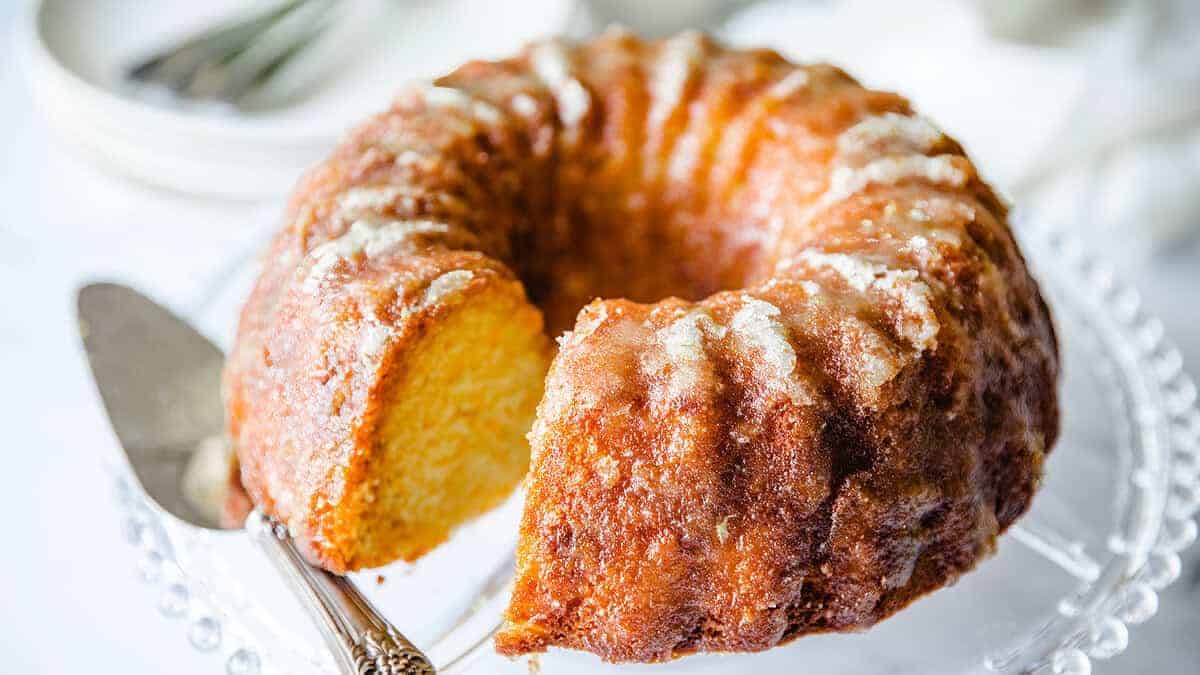 A moist, glazed bundt cake with a golden crust is displayed on a glass cake stand. A slice has been cut, revealing the soft, yellow interior. A cake server rests next to it.