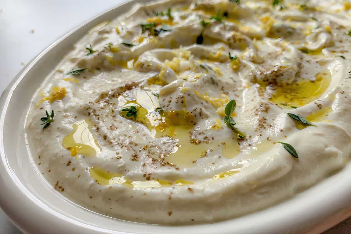 A close-up of a creamy dip garnished with olive oil, ground spices, lemon zest, and fresh herbs on top, served in a white dish.