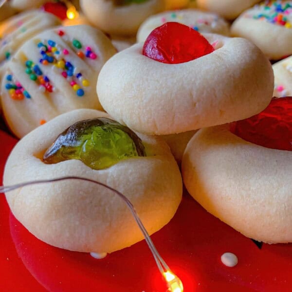 A close-up of assorted cookies with colorful toppings and jelly centers on a red plate, surrounded by string lights.