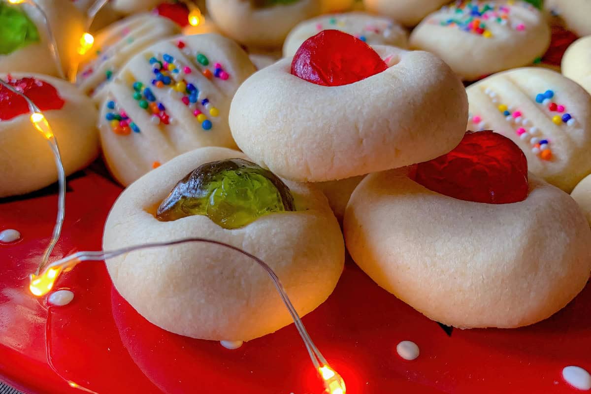A close-up of assorted cookies with colorful toppings and jelly centers on a red plate, surrounded by string lights.