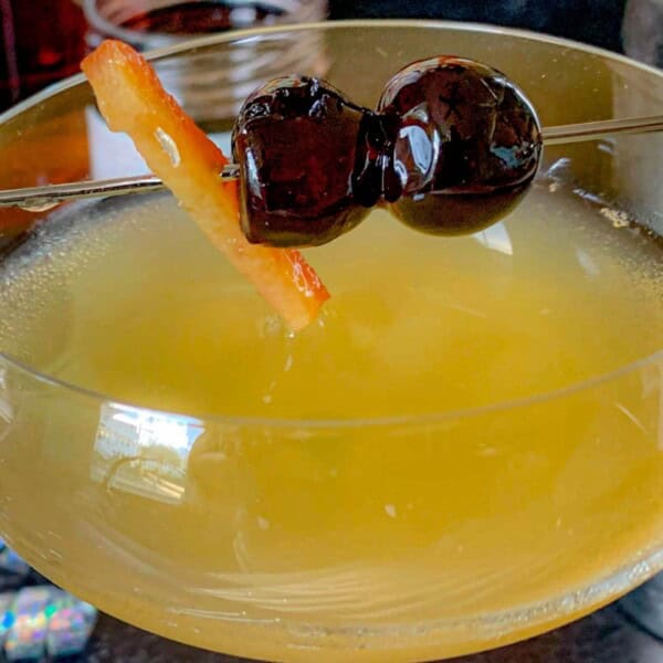 A cocktail glass containing a beverage, adorned with a strip of citrus peel and two dark cherries on a cocktail pick.