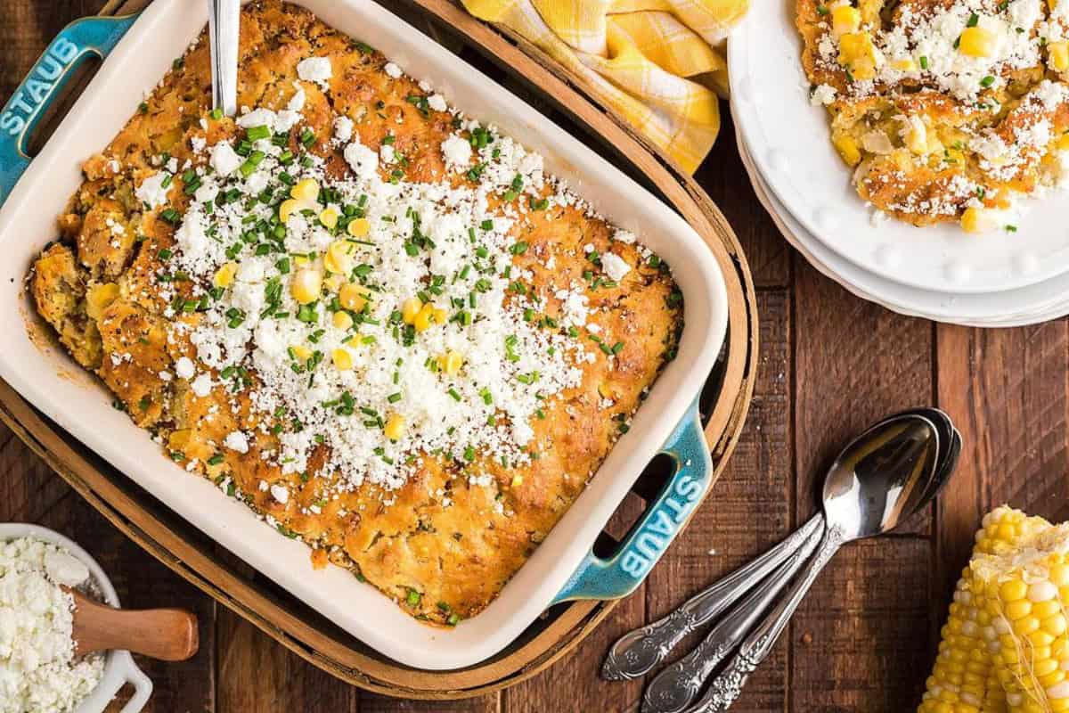 A baked casserole dish containing corn bread topped with crumbled cheese and herbs. Beside it, a plate with a served slice, silverware, and a cob of corn on a wooden table.