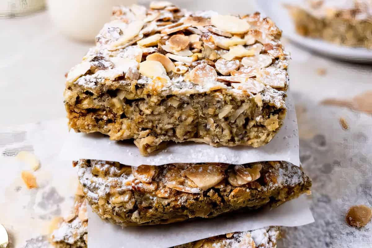 The image shows a stack of oat and almond bars sprinkled with powdered sugar, resting on parchment paper.