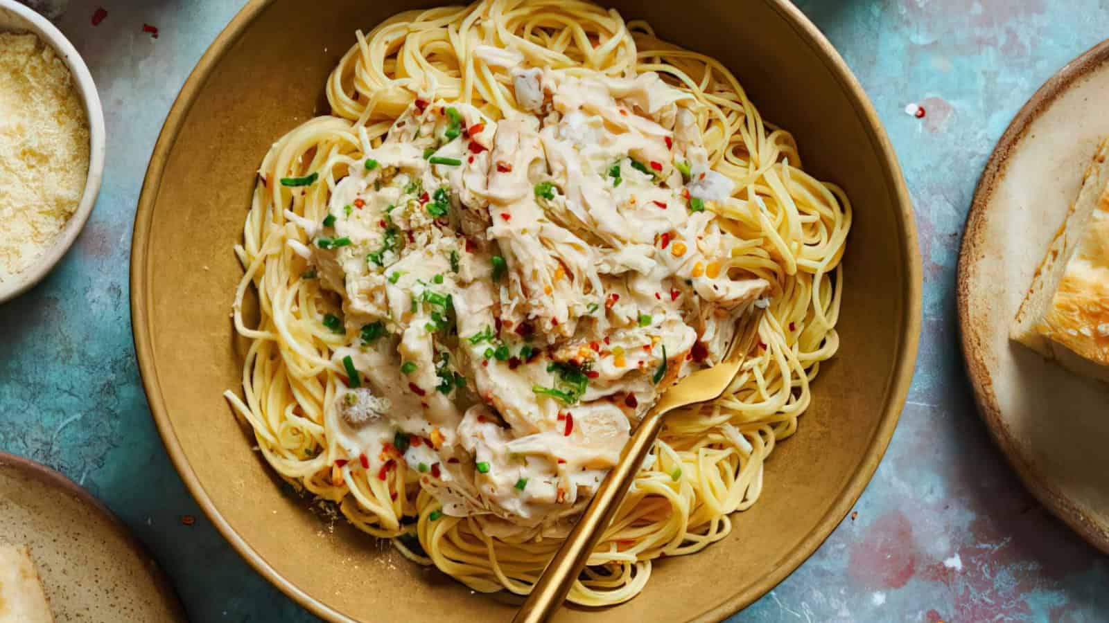 A bowl of spaghetti topped with creamy chicken alfredo sauce, garnished with chopped herbs and red pepper flakes. There is a fork resting in the bowl. Surrounding the bowl are small dishes and a piece of crusty bread.