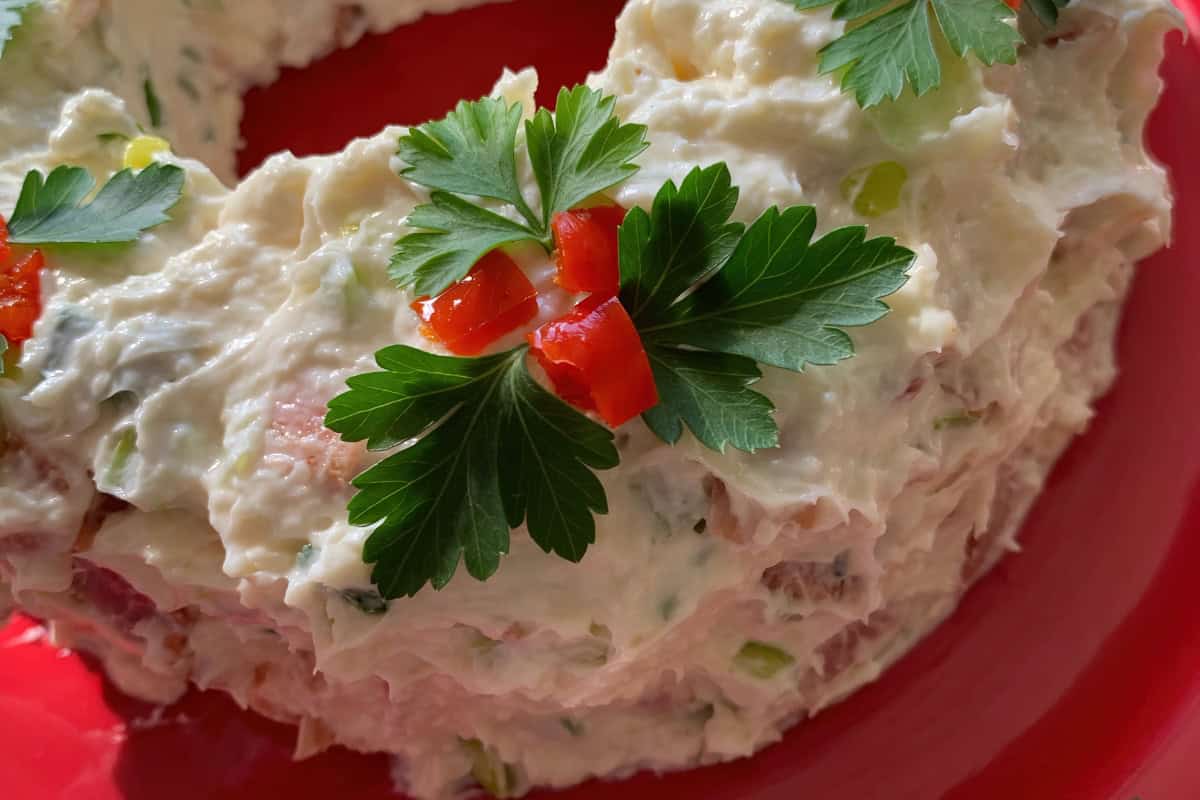 A close-up of a savory salad ring on a red plate, topped with parsley and diced red bell peppers. The dish appears creamy, with visible chunks, possibly containing vegetables or seafood.