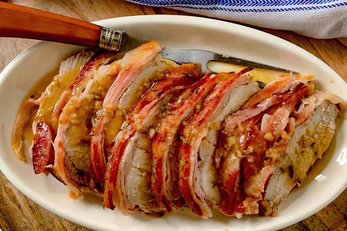 A sliced roasted pork dish wrapped in crispy bacon, placed on a white oval platter. A serving knife rests beside the meat on a wooden table, with a blue and white cloth partially visible in the background.