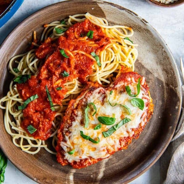 A plate of spaghetti with tomato sauce and chicken parmesan, garnished with fresh basil, next to a fork and knife on a napkin.
