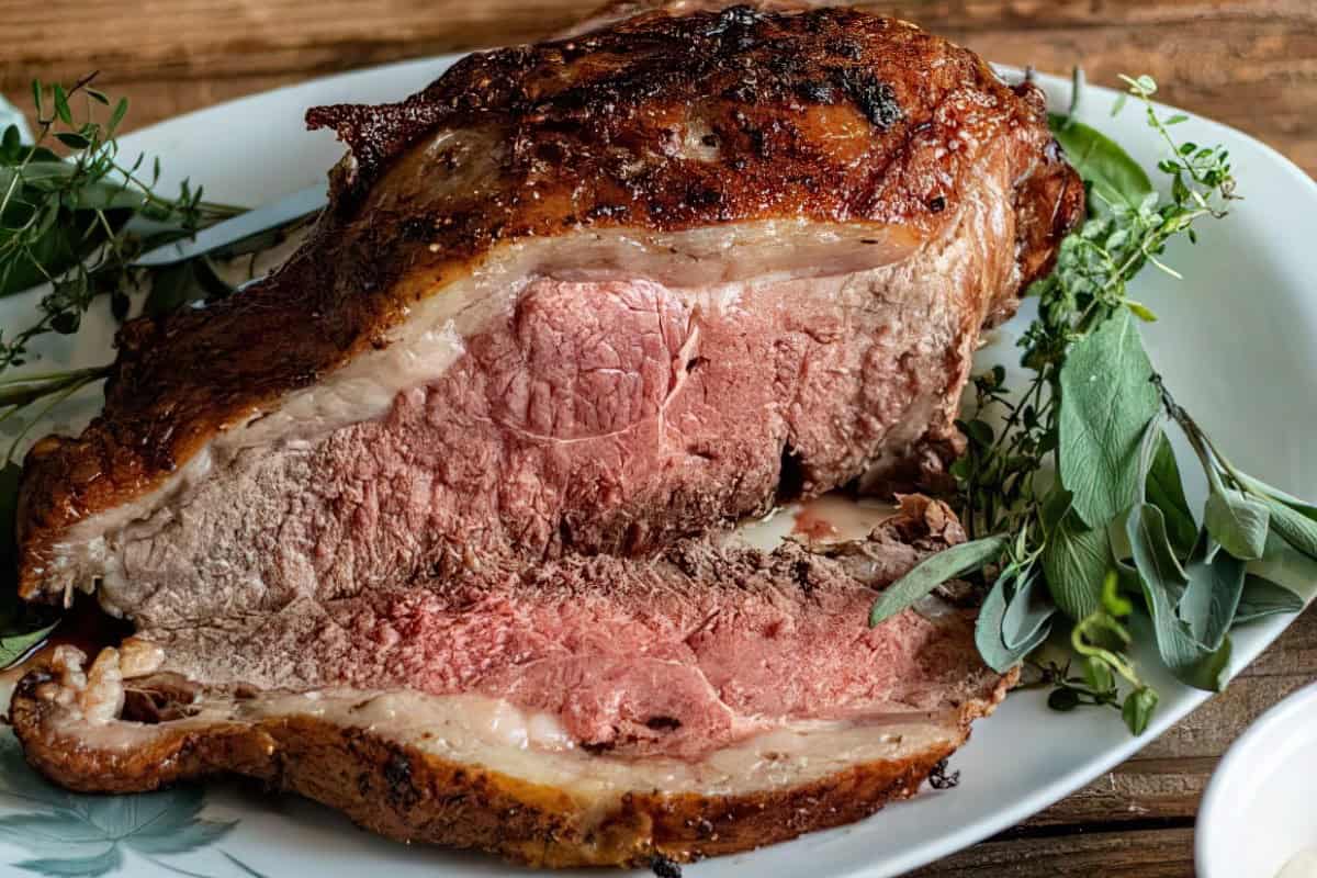 A succulent roast beef with a browned, crispy exterior and pink, juicy interior is sliced open on a white plate. Fresh herbs, including sage and thyme, garnish the plate, adding a touch of green.