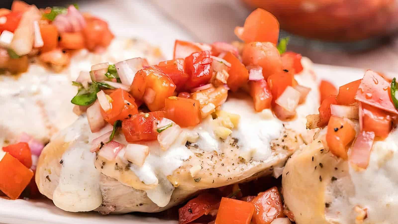 Close-up of baked chicken breasts topped with melted cheese and a colorful bruschetta mix of diced tomatoes, onions, and herbs. The dish is arranged on a white plate with a blurred background.