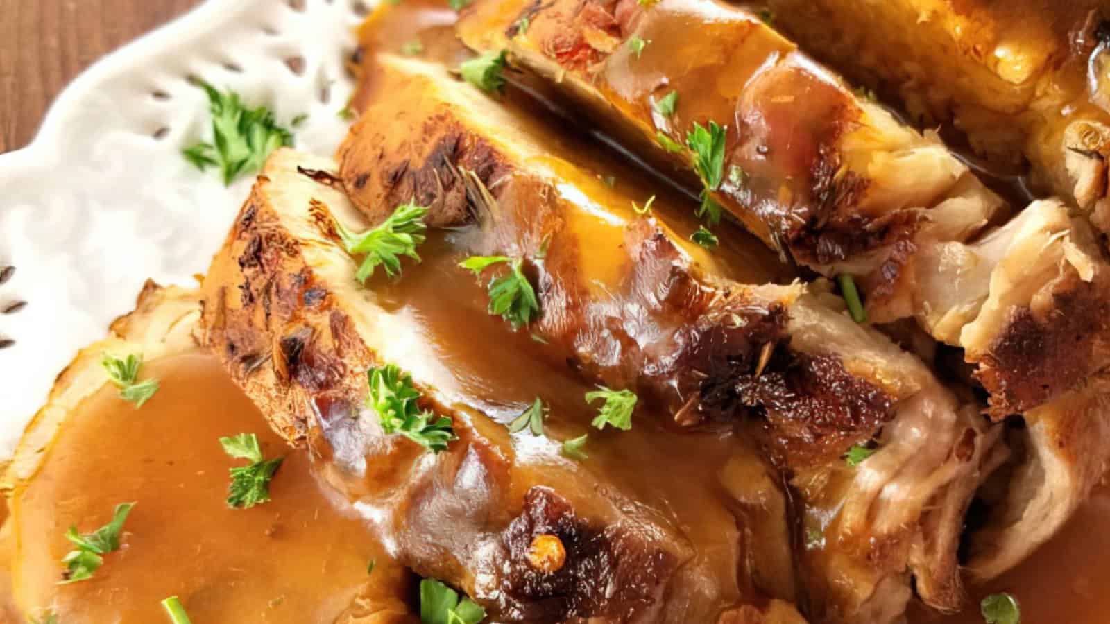 Close-up of a roasted meat dish, sliced and topped with a glossy brown gravy. Garnished with fresh parsley, the dish is served on a white plate.