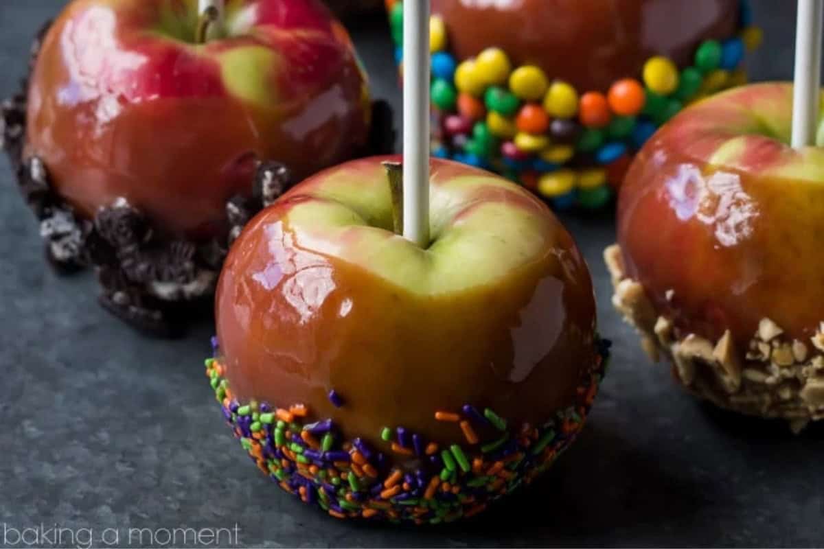 Close-up of caramel apples with decorative toppings. One apple has colorful sprinkles, another has bits of chocolate cookies, and a third has chopped nuts. A variety of candies adorn the apples, which are set on a dark surface.