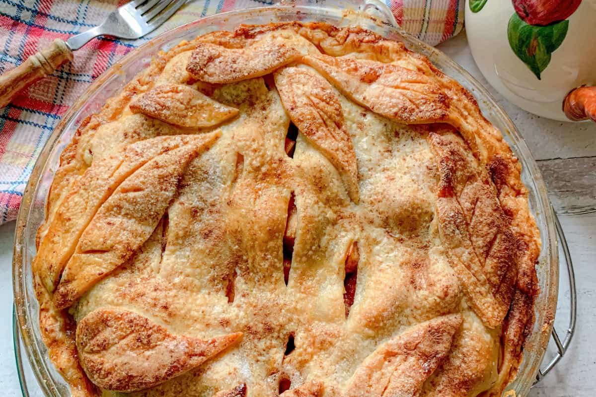 A baked pie with a golden-brown crust and decorative leaf-shaped cutouts is on a table near a fork and a patterned cloth.