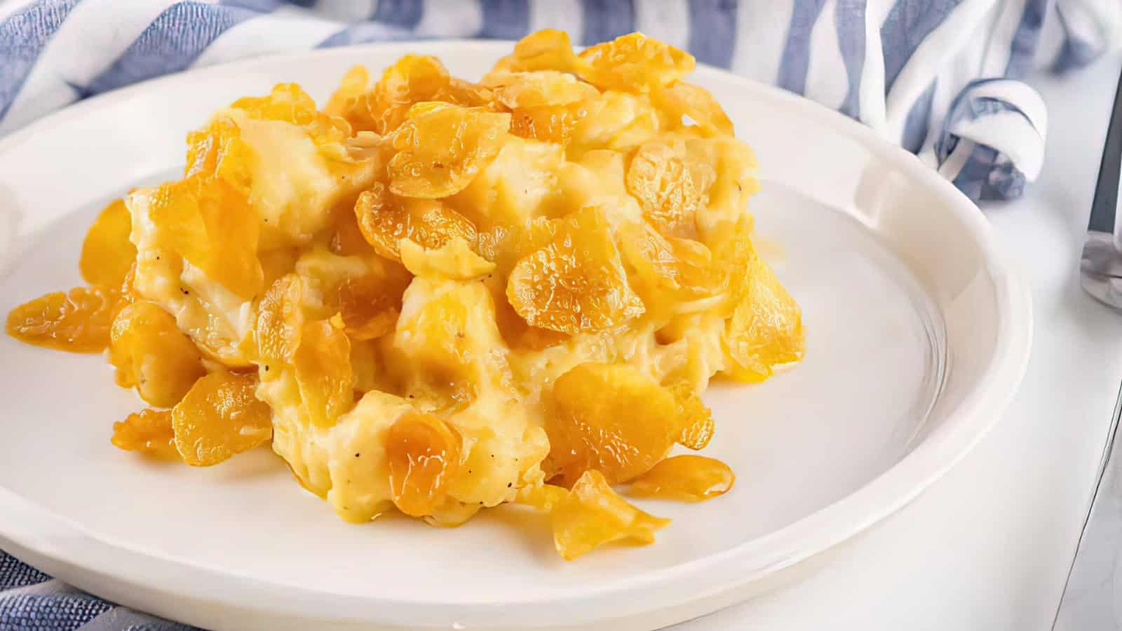 A plate of creamy cheesy potatoes topped with golden cornflakes, set on a white dish with a striped cloth in the background. The dish appears rich and comforting.