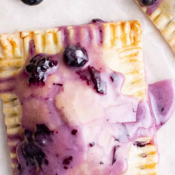 Rectangular pastry with purple blueberry glaze and whole blueberries, surrounded by other pastries and loose blueberries on a light surface.