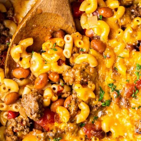 A skillet with cooked macaroni, ground meat, beans, tomatoes, melted cheese, and herbs; a wooden spoon stirs the mixture.