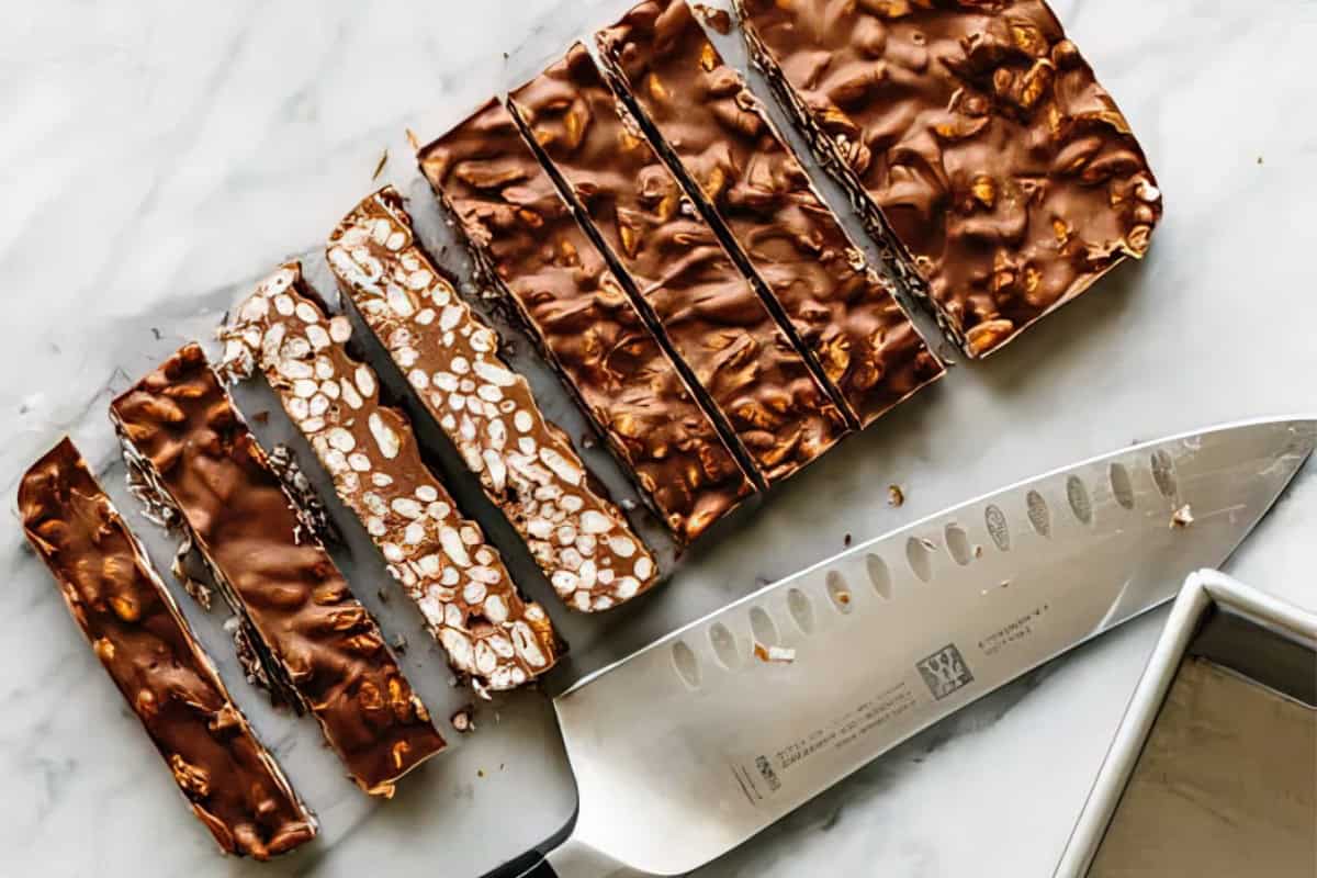 Sliced chocolate bars with a nutty texture are displayed on a marble surface. A knife with a silver blade is placed nearby, along with a metal baking pan in the corner.