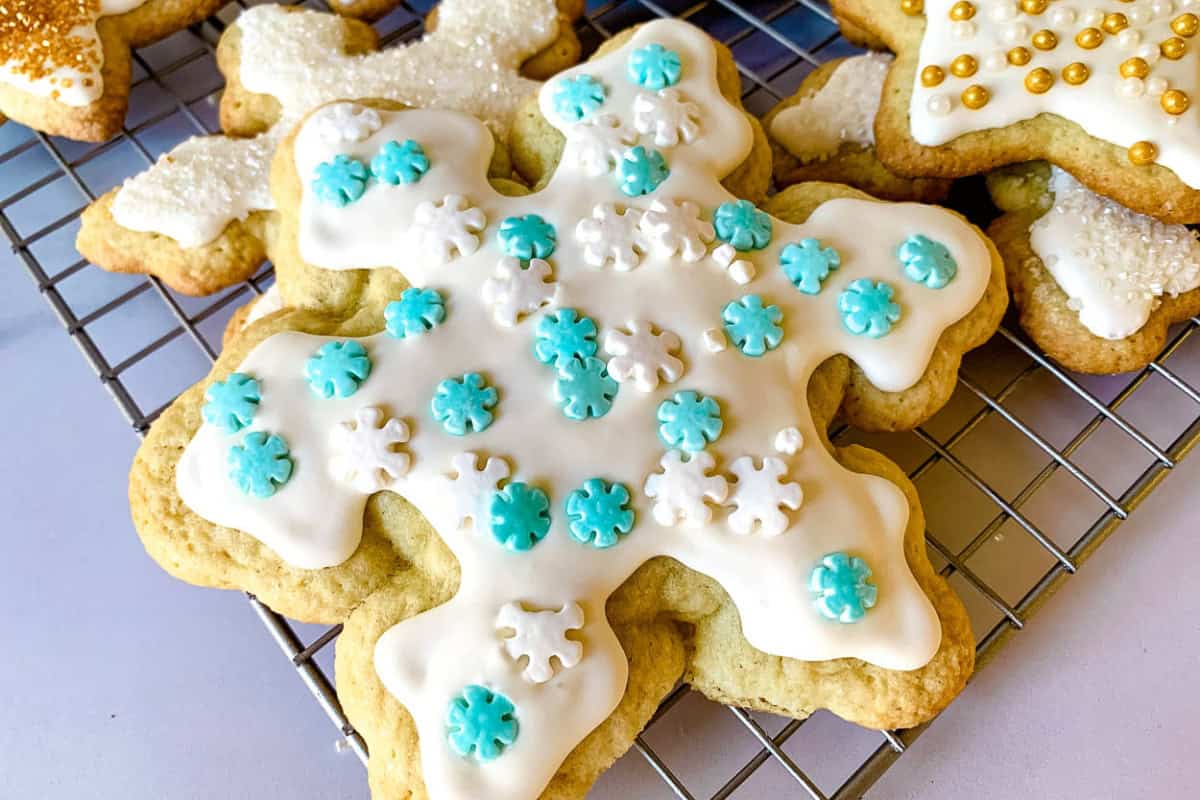 A snowflake-shaped cookie is decorated with white icing and topped with blue and white snowflake accents while cooling on a rack.