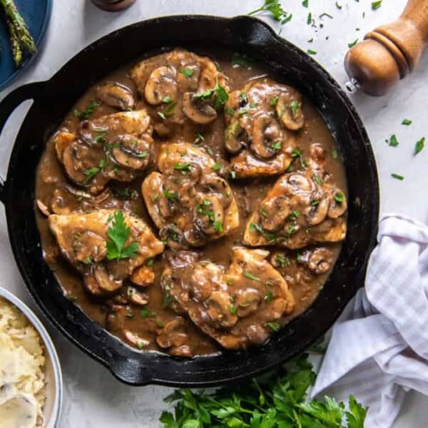 A cast iron skillet of chicken in mushroom sauce sits next to a plate with asparagus, mashed potatoes, parsley, and a pepper grinder.