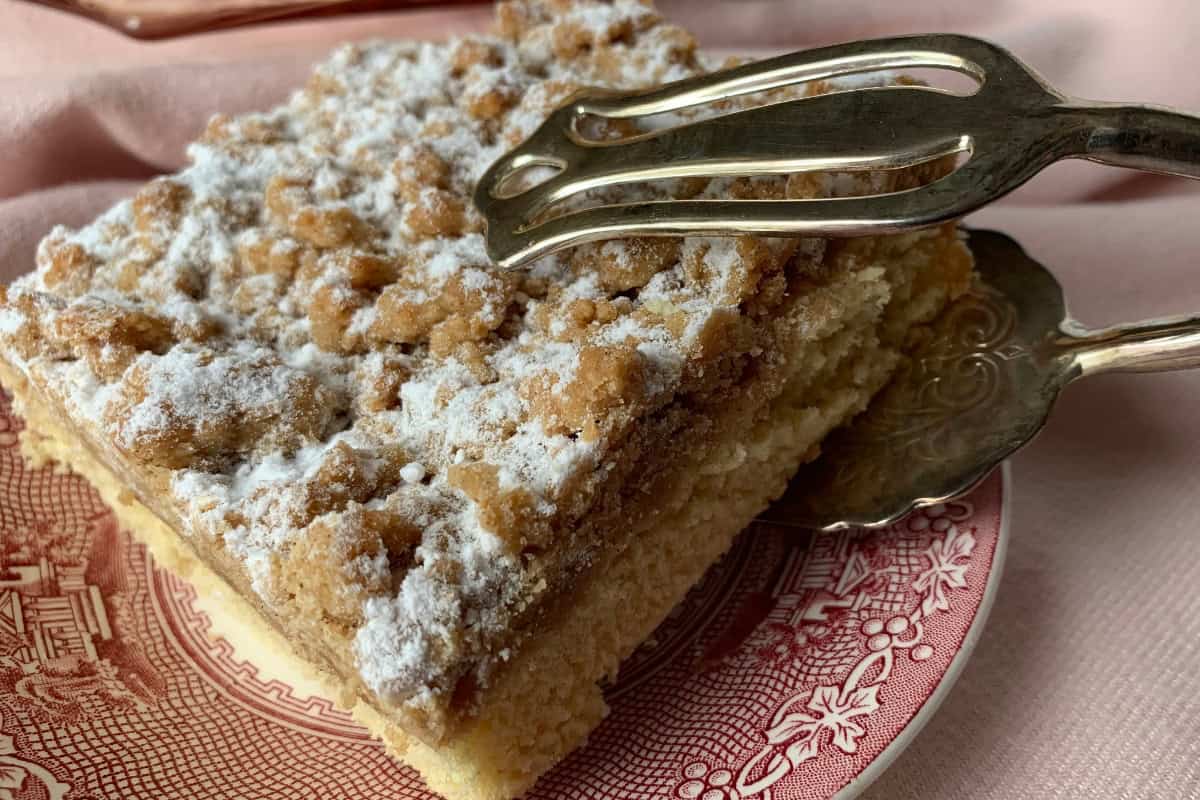 A piece of crumble cake with powdered sugar on a decorative pink plate. A silver cake server partially slides under the slice, resting on a soft pink fabric.