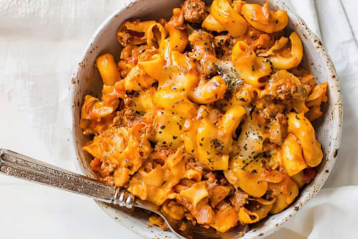 A bowl of cheesy pasta mixed with ground beef and herbs, accompanied by a fork.
