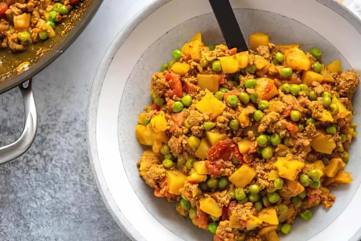 A white bowl filled with a colorful mixture of cooked ground meat, potatoes, peas, and tomatoes. A serving spoon is placed in the bowl. Another pan with the same food is partially visible on the side.
