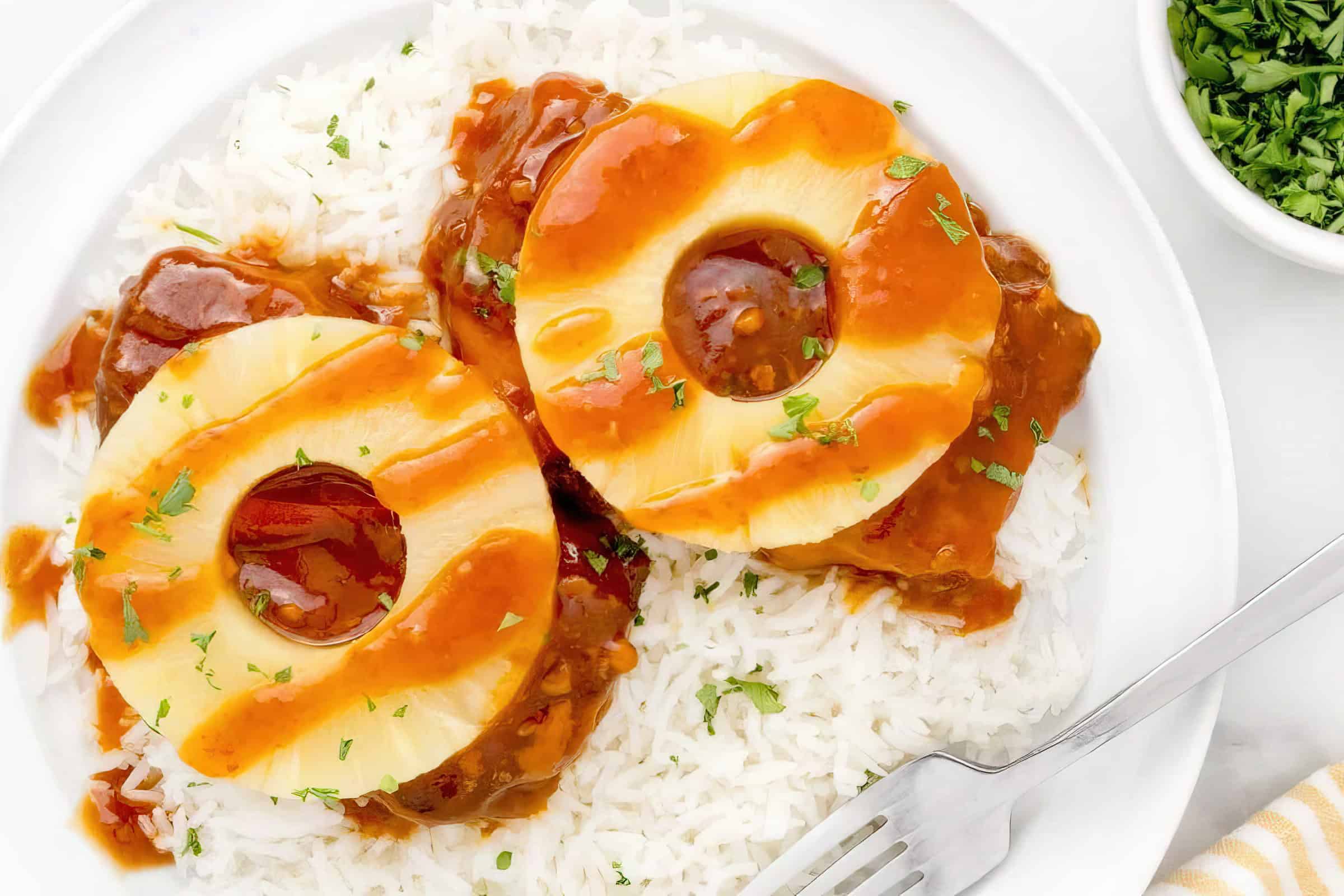 A plate of white rice topped with two slices of pineapple and meat in a rich brown sauce, garnished with herbs. A fork is placed on the edge of the plate, with a small dish of chopped herbs nearby.