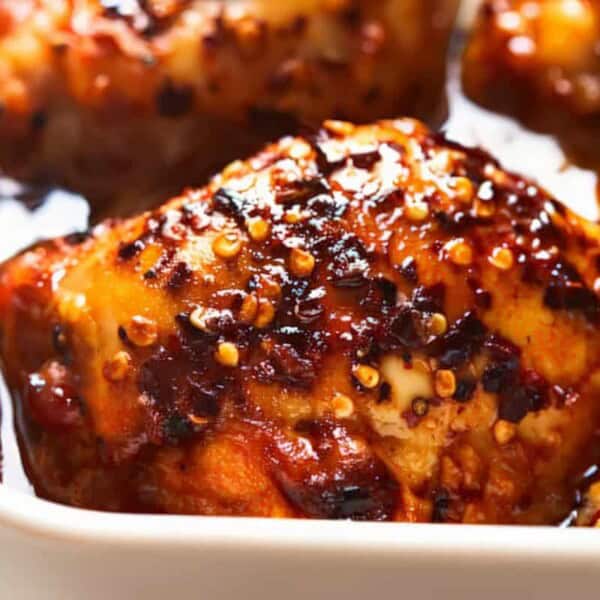 Close-up of glazed baked chicken thighs topped with visible spices and seeds, arranged in a white baking dish.