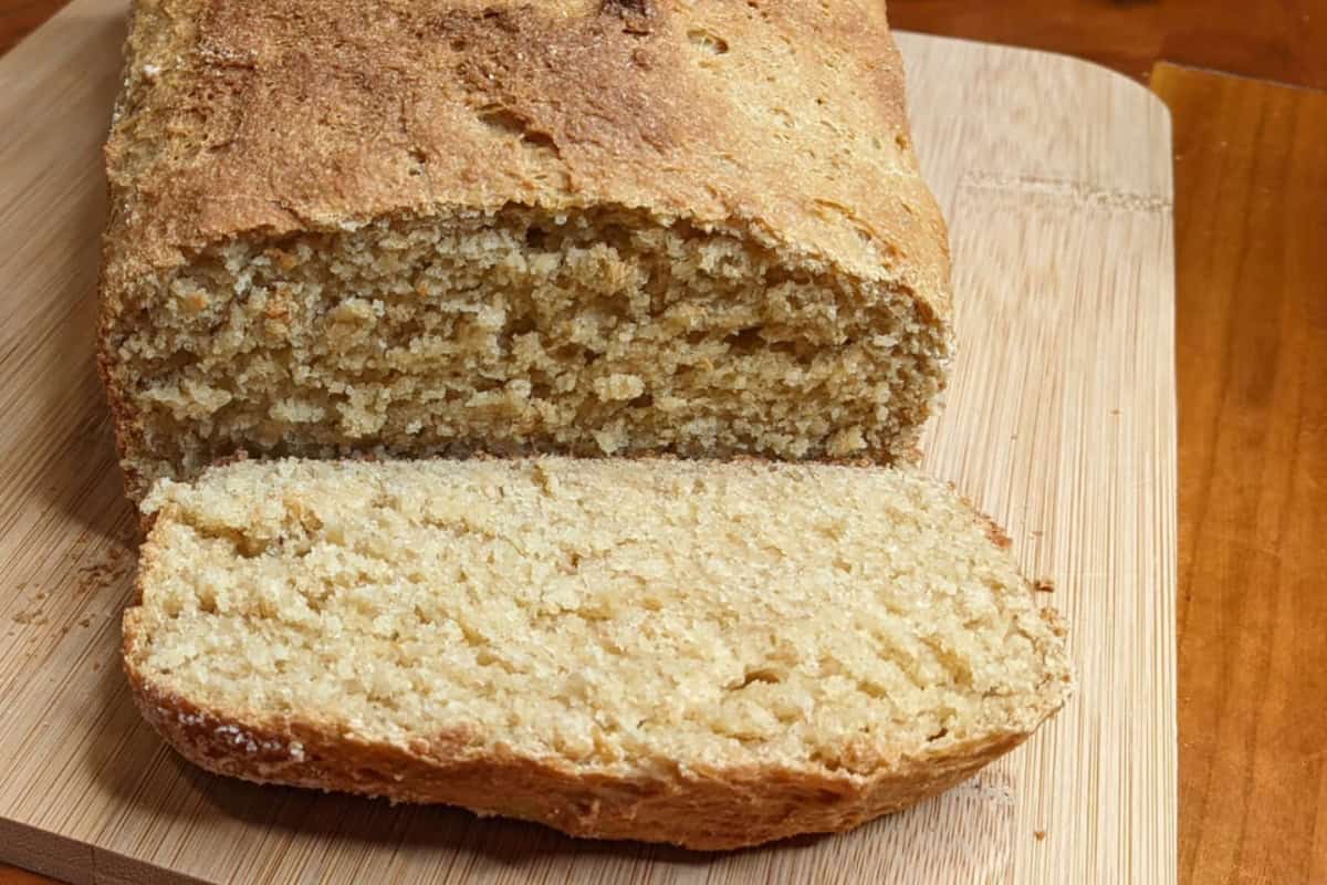 A loaf of freshly baked bread with a rough crust and soft, crumbly texture sits on a wooden cutting board. One slice is cut and placed in front, revealing its light, fluffy interior. The background is a warm, brown wood surface.