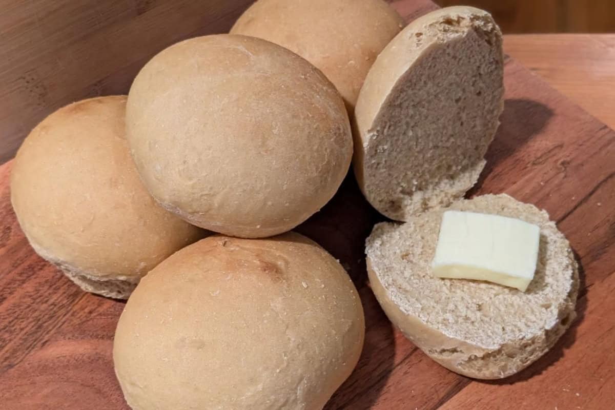 A wooden board with five freshly baked bread rolls. One roll is cut open, displaying a pat of butter placed on the bottom half. The surface has a warm, golden brown texture.