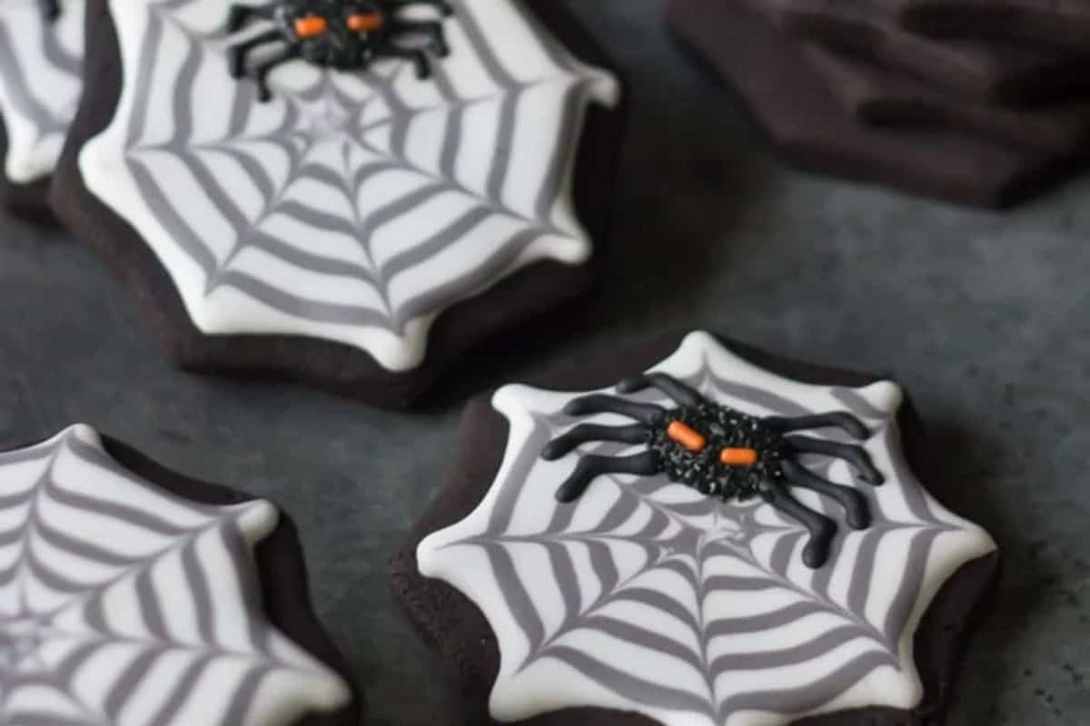 Halloween-themed cookies shaped like spider webs, decorated with icing and featuring small spiders made of icing on top. The cookies are arranged on a dark surface.