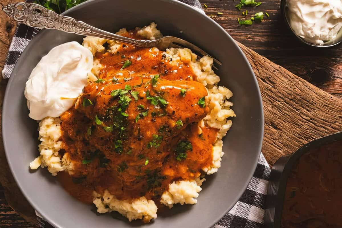 A bowl of creamy mashed potatoes topped with a piece of chicken covered in a rich, spiced paprika sauce. It's garnished with fresh herbs and served with a dollop of sour cream, accompanied by a decorative fork on the side.