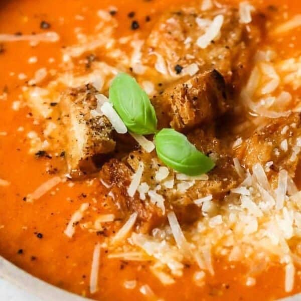 Bowl of tomato soup topped with croutons, grated cheese, ground pepper, and basil leaves; spoon on the side.