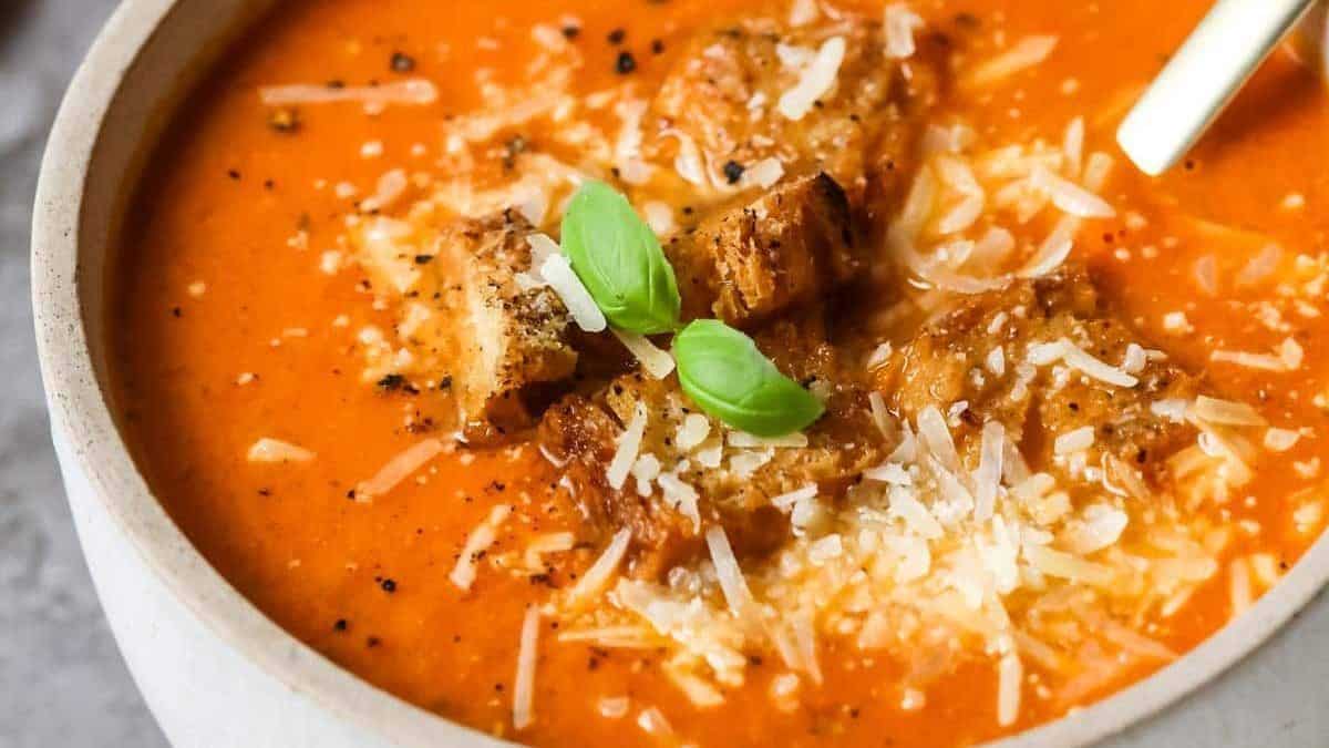 Bowl of tomato soup topped with croutons, grated cheese, ground pepper, and basil leaves; spoon on the side.
