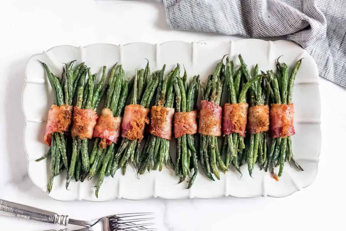 Green beans wrapped in bacon, arranged in neat bundles on a white serving platter with a gray napkin and forks beside it.