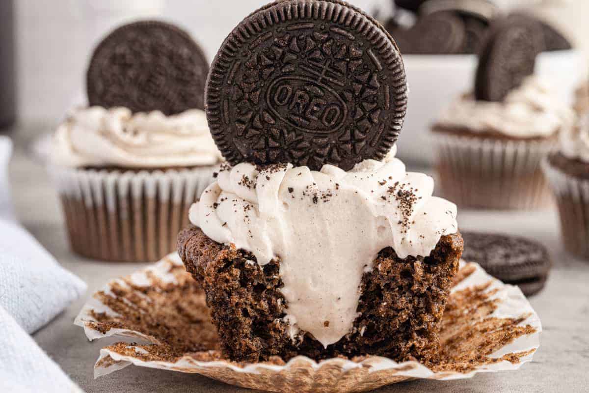 A chocolate cupcake with cookies and cream frosting, topped with an Oreo, sits in an open wrapper with a bite showing creamy filling.