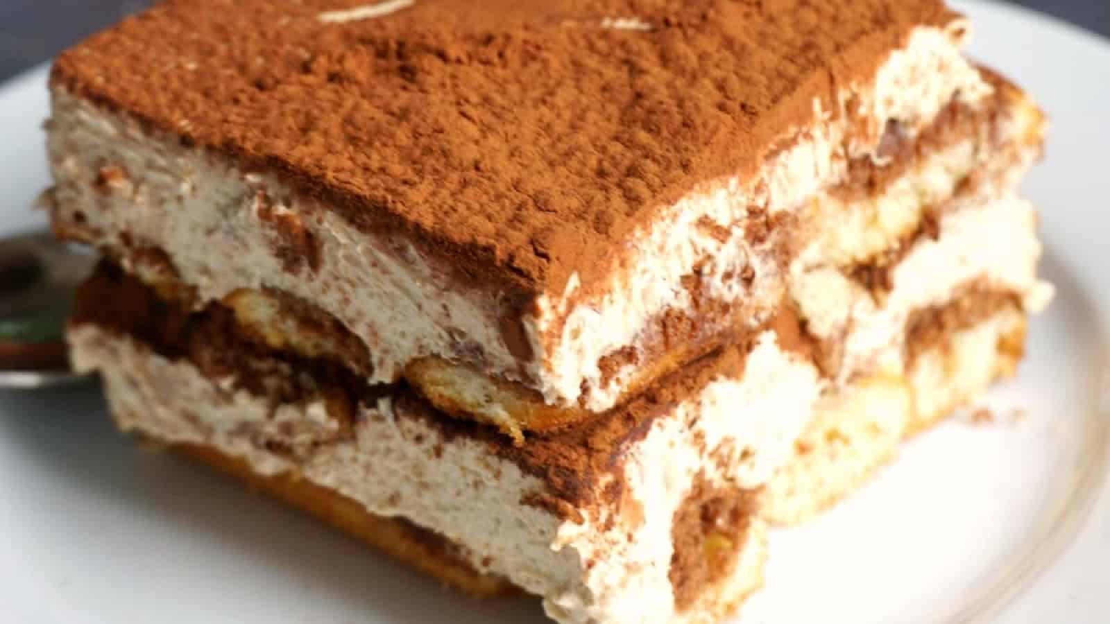 A close-up of a slice of tiramisu on a white plate. The dessert has layers of creamy mascarpone filling and coffee-soaked ladyfingers, topped with a dusting of cocoa powder. A spoon is partially visible beside the tiramisu.