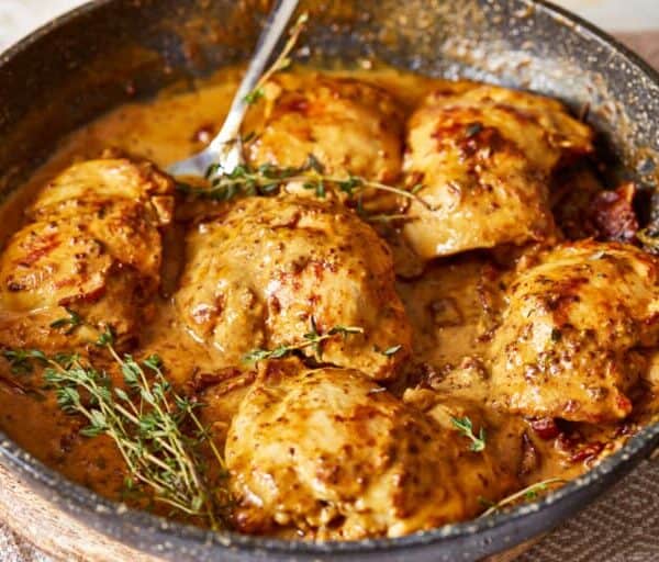 Chicken thighs in creamy mustard sauce with fresh thyme garnish, cooking in a black skillet.