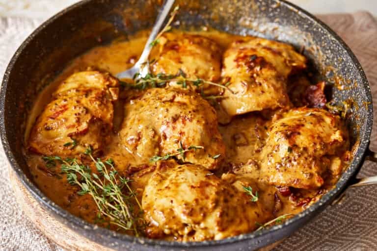 Chicken thighs in creamy mustard sauce with fresh thyme garnish, cooking in a black skillet.