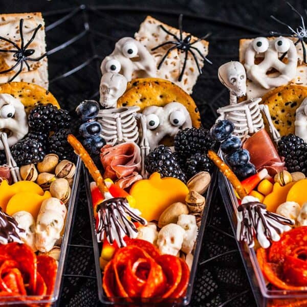 Three snack cups with crackers, cheese, meats, berries, nuts, skeleton and ghost picks, and plastic spider toppers.